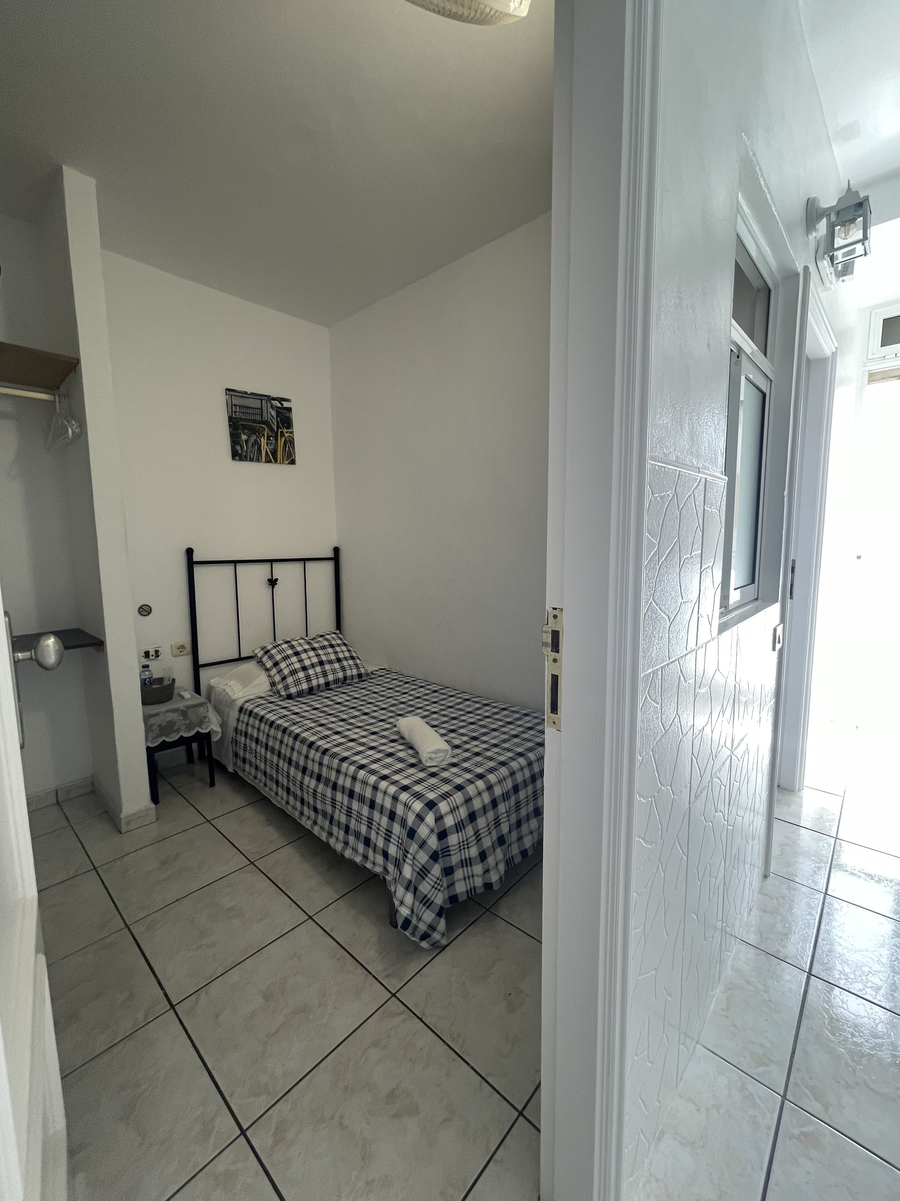 A bedroom with a bed, a nightstand, a small table, a wall-mounted light, and a door leading to another room.