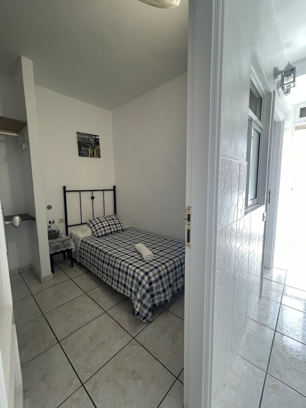 A bedroom with a bed, a nightstand, a small table, a wall-mounted light, and a door leading to another room. - Pension Casa Blanca (Arona)