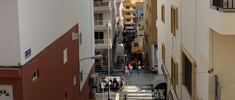 A narrow street with buildings on either side, a staircase leading to a higher level, and a view of the sea in the distance.