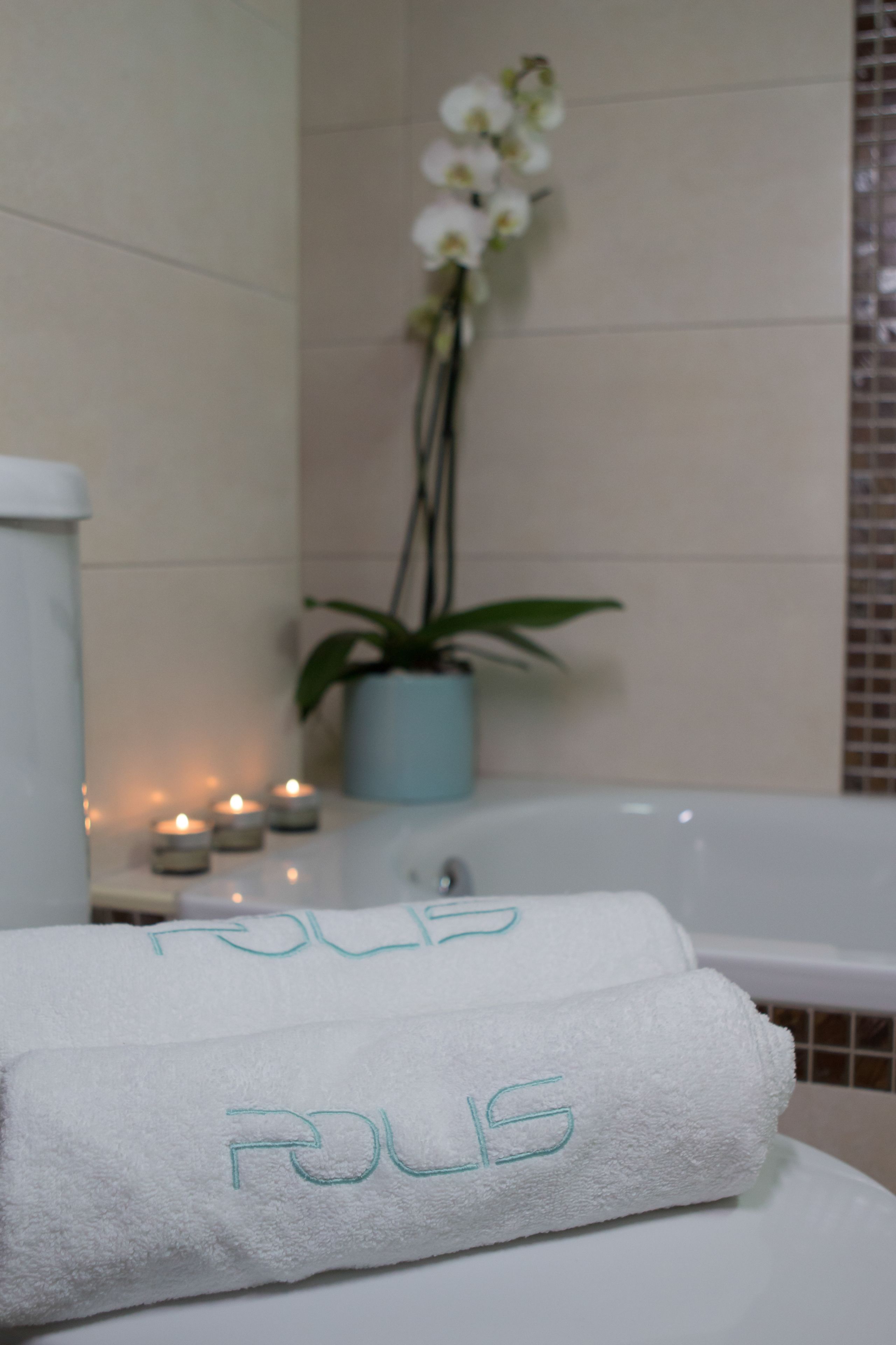 A bathroom with a bathtub, two towels with embroidered text, a potted plant, and candles.