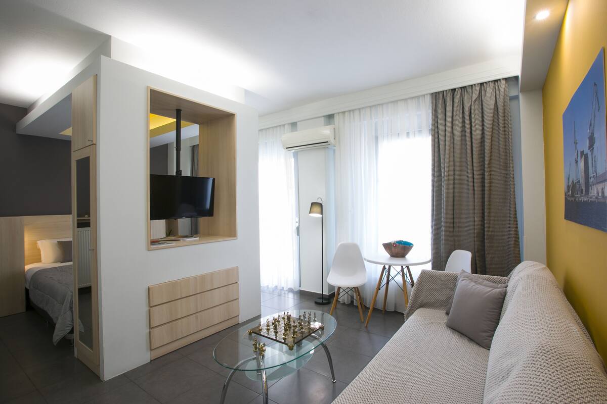 A modern living room with a glass coffee table, a sofa, a TV mounted on a wall, and a dining area with a chair and table.
