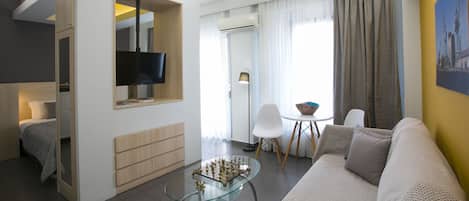 Uma sala de estar moderna com uma mesa de centro de vidro, um sofá, uma TV fixada na parede e um espaço de jantar com uma cadeira e mesa.