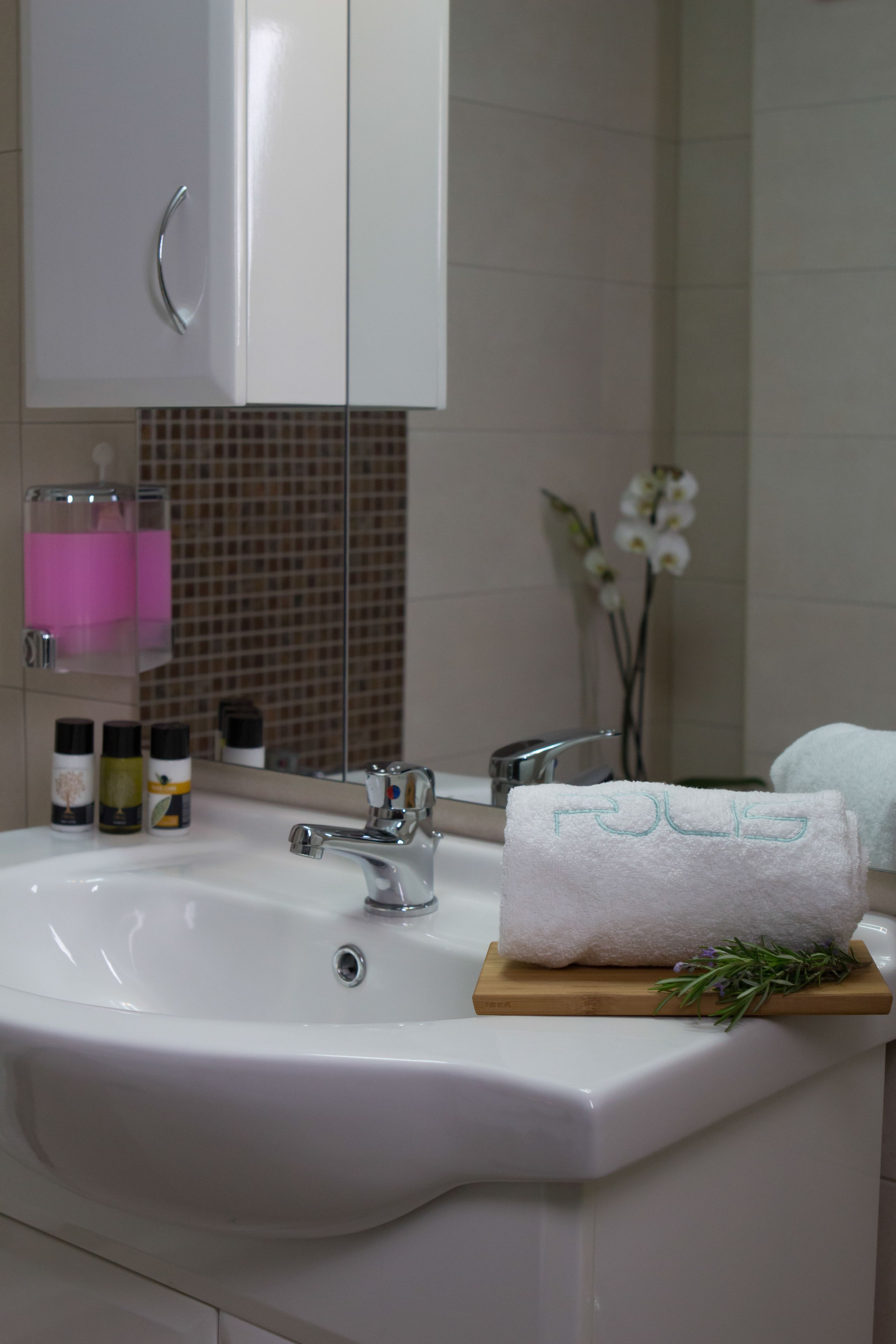 A bathroom with a white sink, a mirror, a towel rack, and a small wooden tray with a towel and a sprig of rosemary.