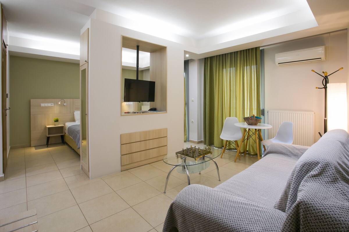 A modern hotel room with a bed, a glass coffee table, a TV, and a seating area with chairs and a table.