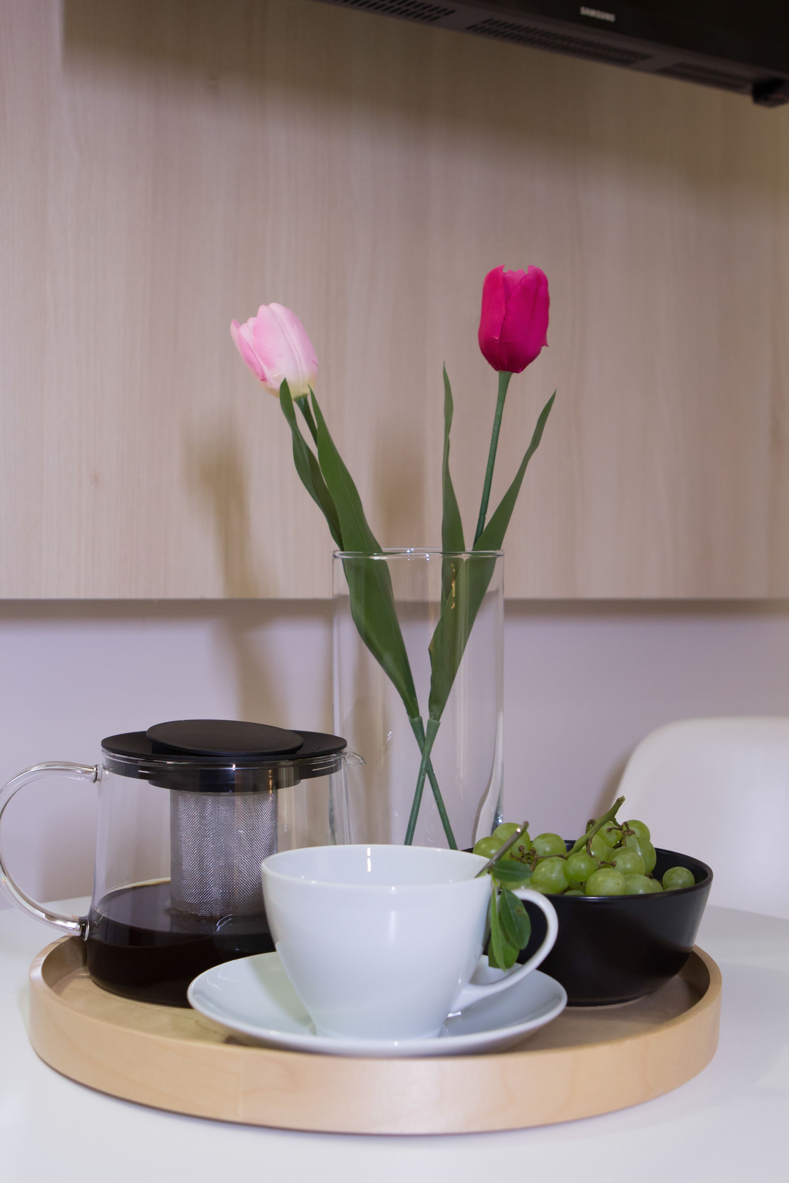A clear glass vase with two tulips, a coffee maker, a white cup, and a bowl of green grapes on a wooden tray.