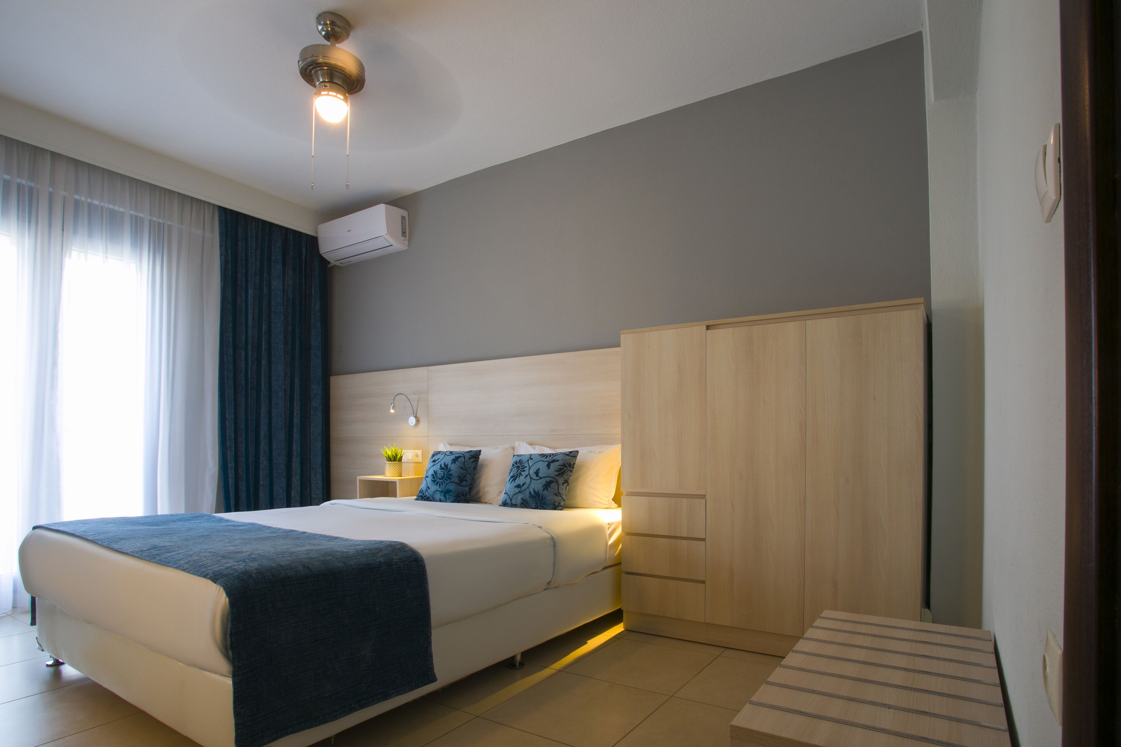 Una habitación de hotel moderna con una cama grande, un armario de madera, una mesita de noche con lámpara y un equipo de aire acondicionado en la pared.