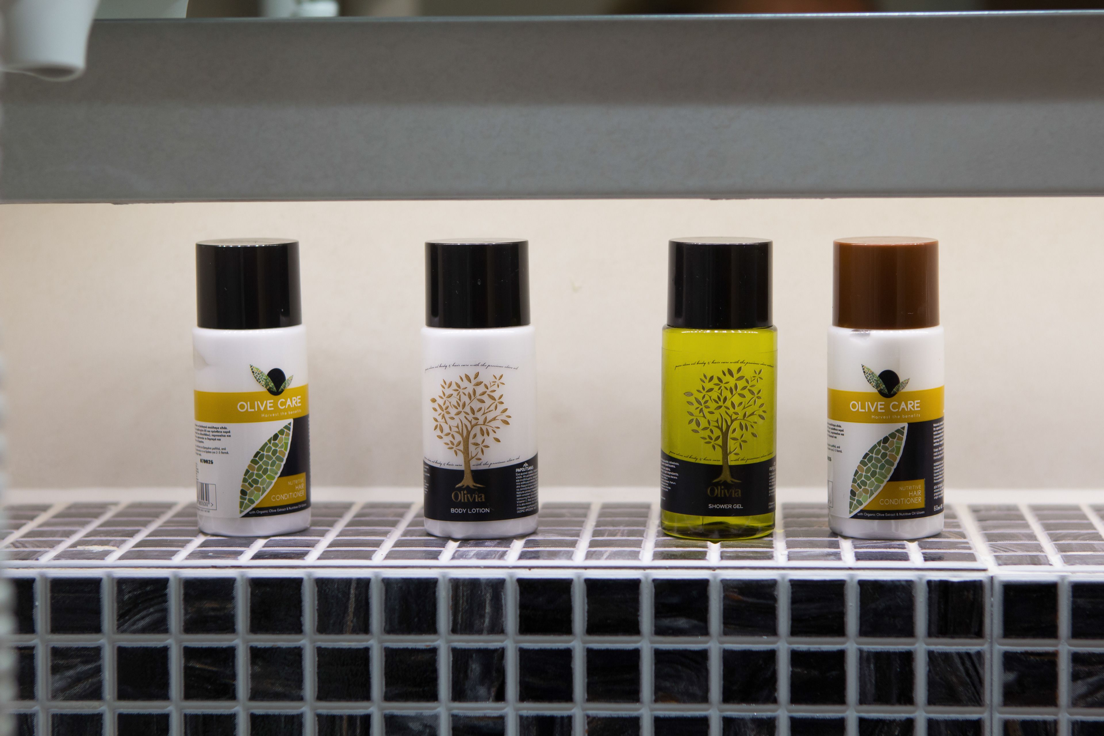 Four bottles of Olive Care body lotion and shower gel on a tiled shelf.