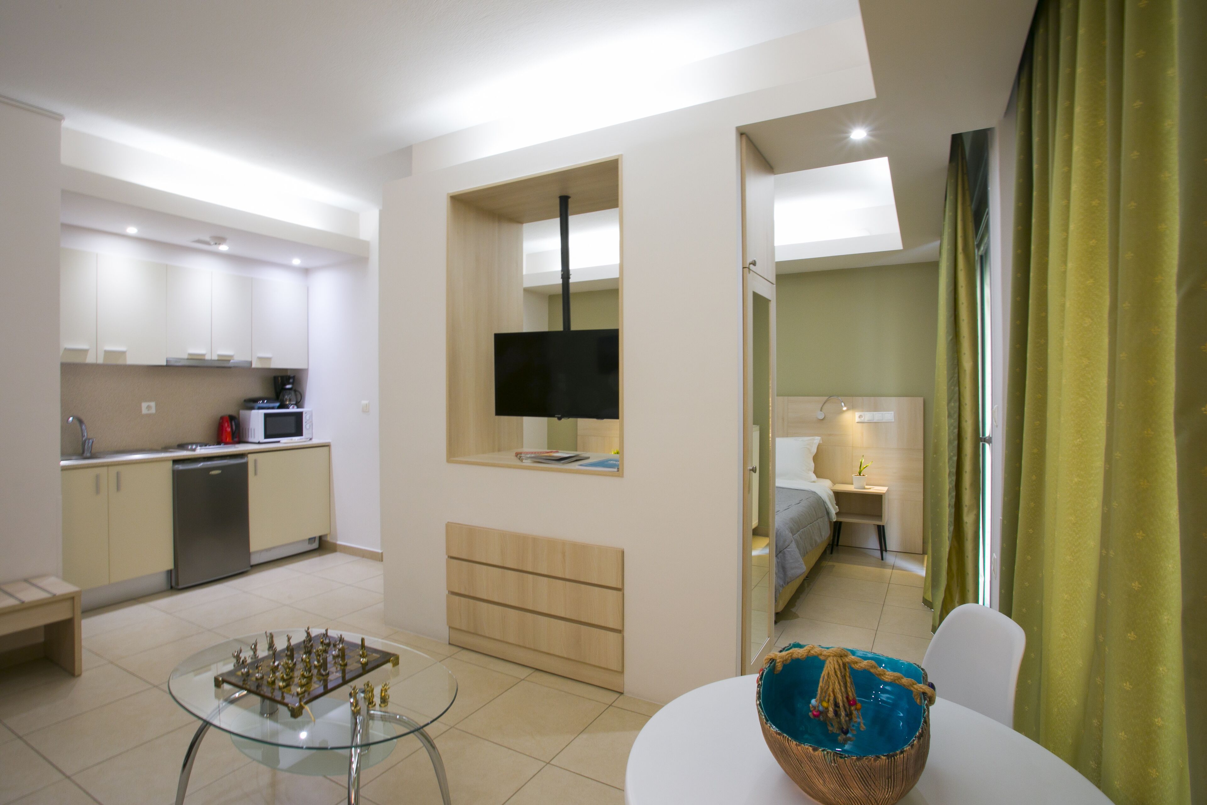 A compact hotel room with a kitchenette, a TV mounted on a wall, a bed, and a small table with a basket.