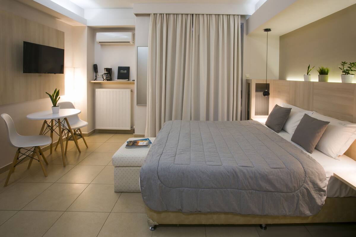 A modern hotel room with a large bed, a desk with a chair, a small table with a plant, and a TV mounted on the wall.