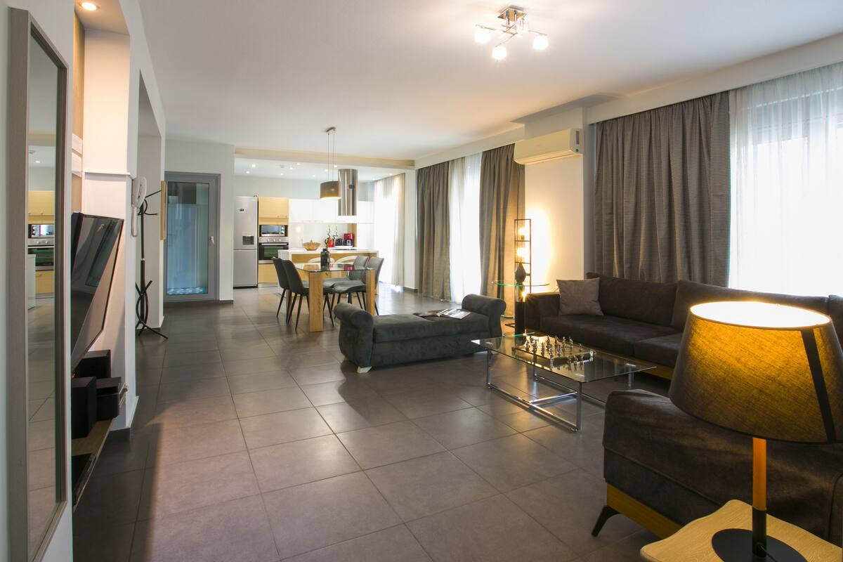 A modern living room with a sofa, a coffee table, and a lamp. There is a dining area in the background with chairs and a table.