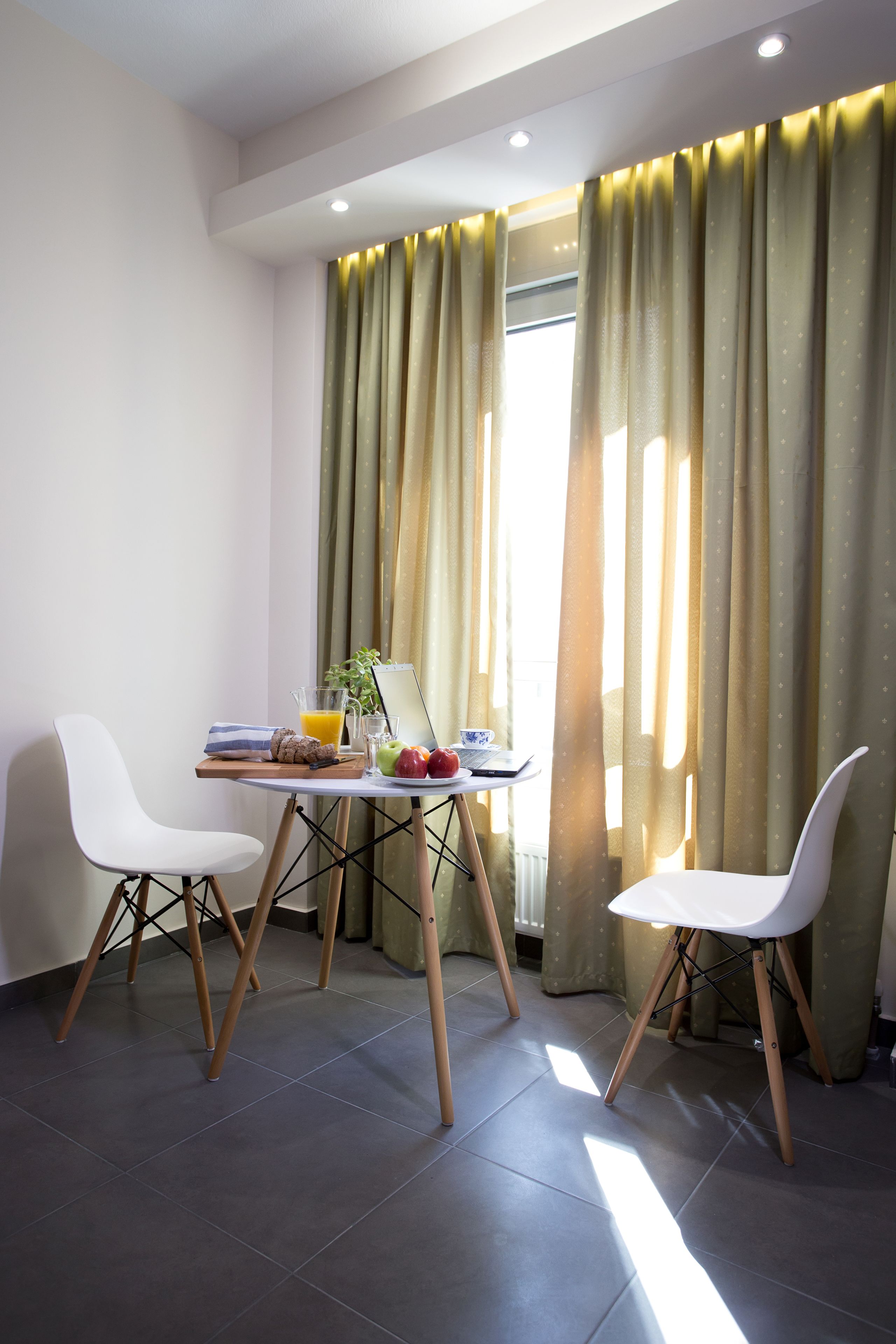 A dining area with a table set for two, a fruit bowl, and a window with curtains.