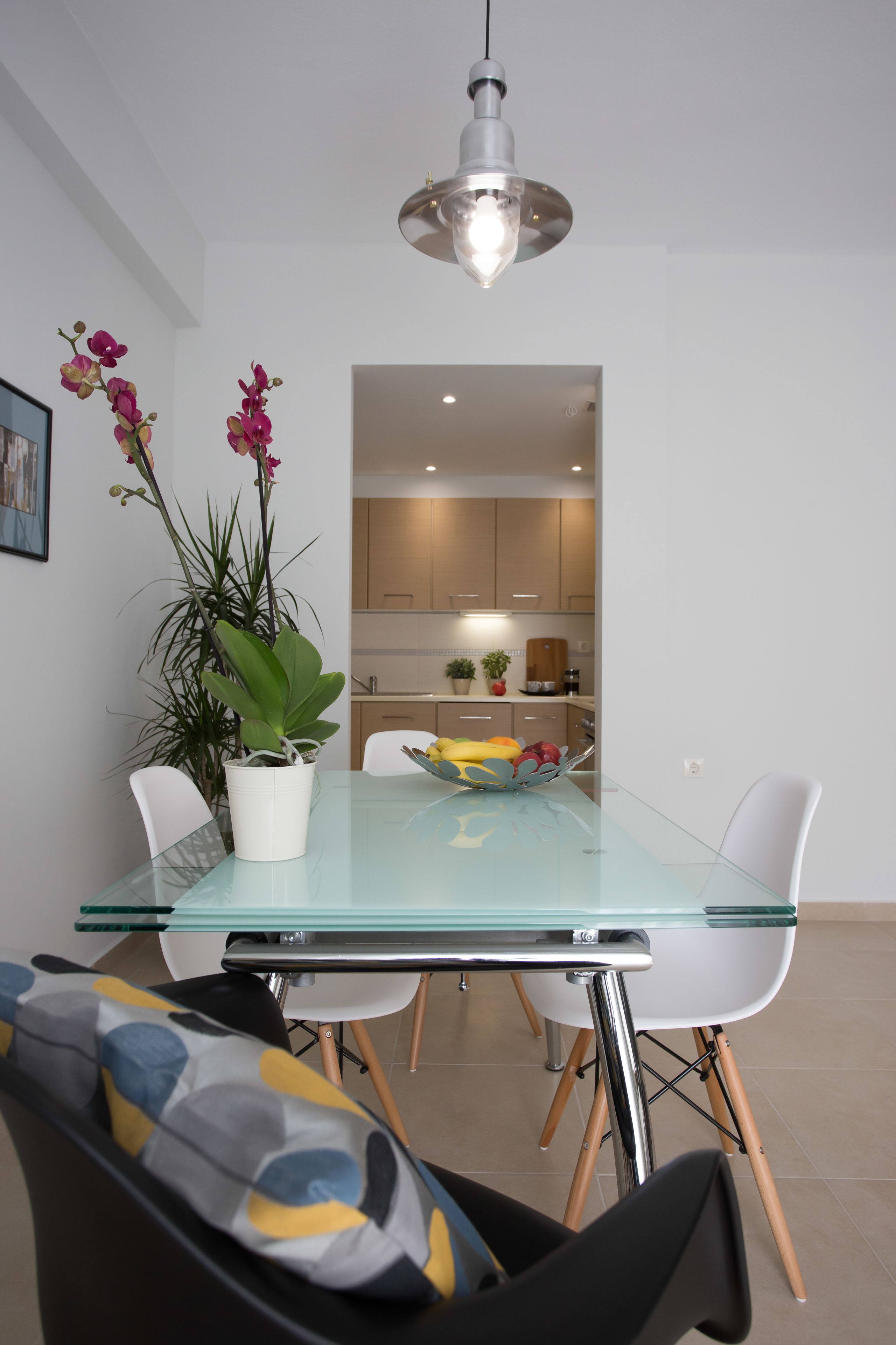 Un espace repas moderne avec une table en verre, des chaises blanches, une plante en pot et un luminaire suspendu.