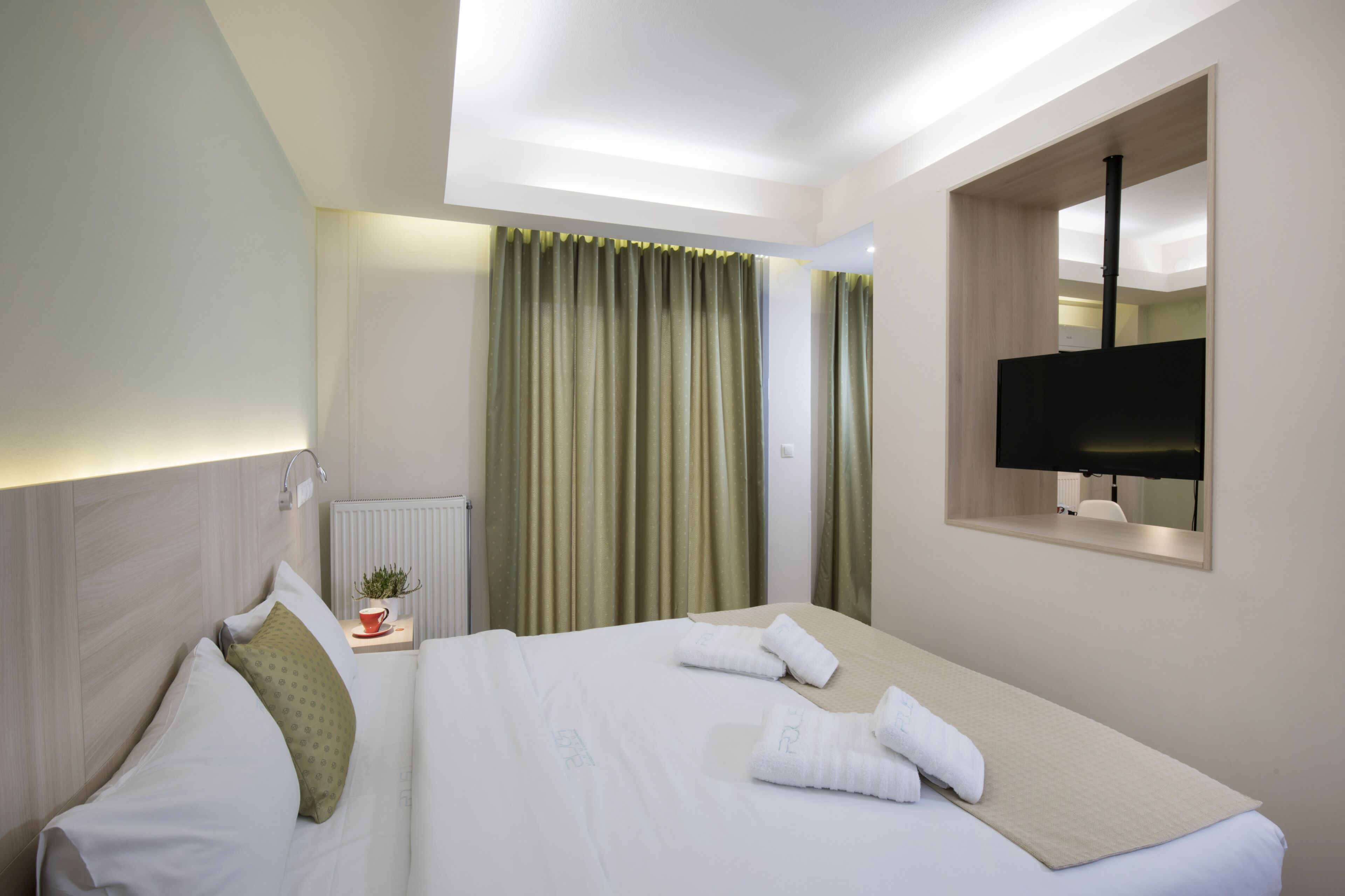 A hotel room with a neatly made bed, a small table with a cup and a plant, a window with curtains, and a TV mounted on the wall.