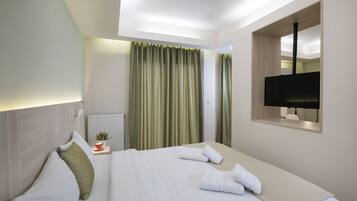 Una habitación de hotel con una cama bien hecha, una mesita con una taza y una planta, una ventana con cortinas y un televisor montado en la pared.