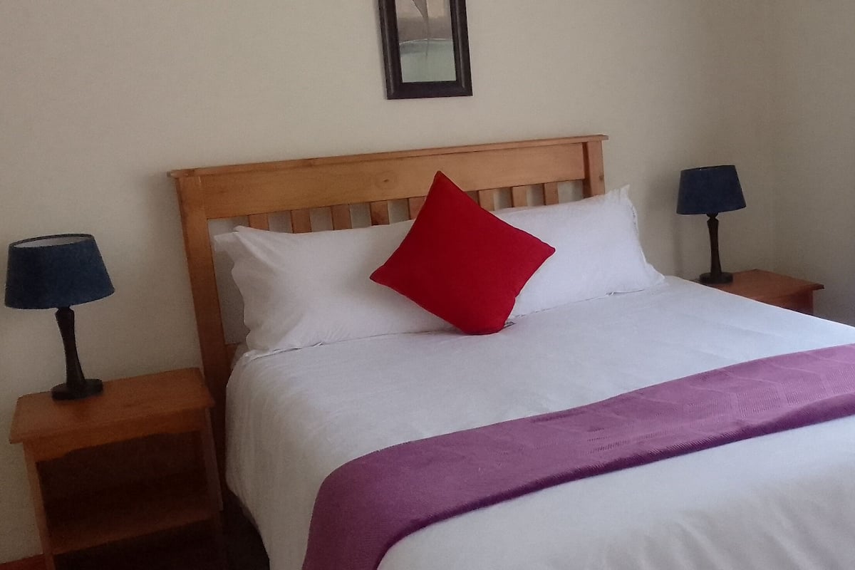 A neatly made bed with a wooden headboard, white bedding, a red pillow, and a purple throw. There are two bedside tables with lamps on either side of the bed.