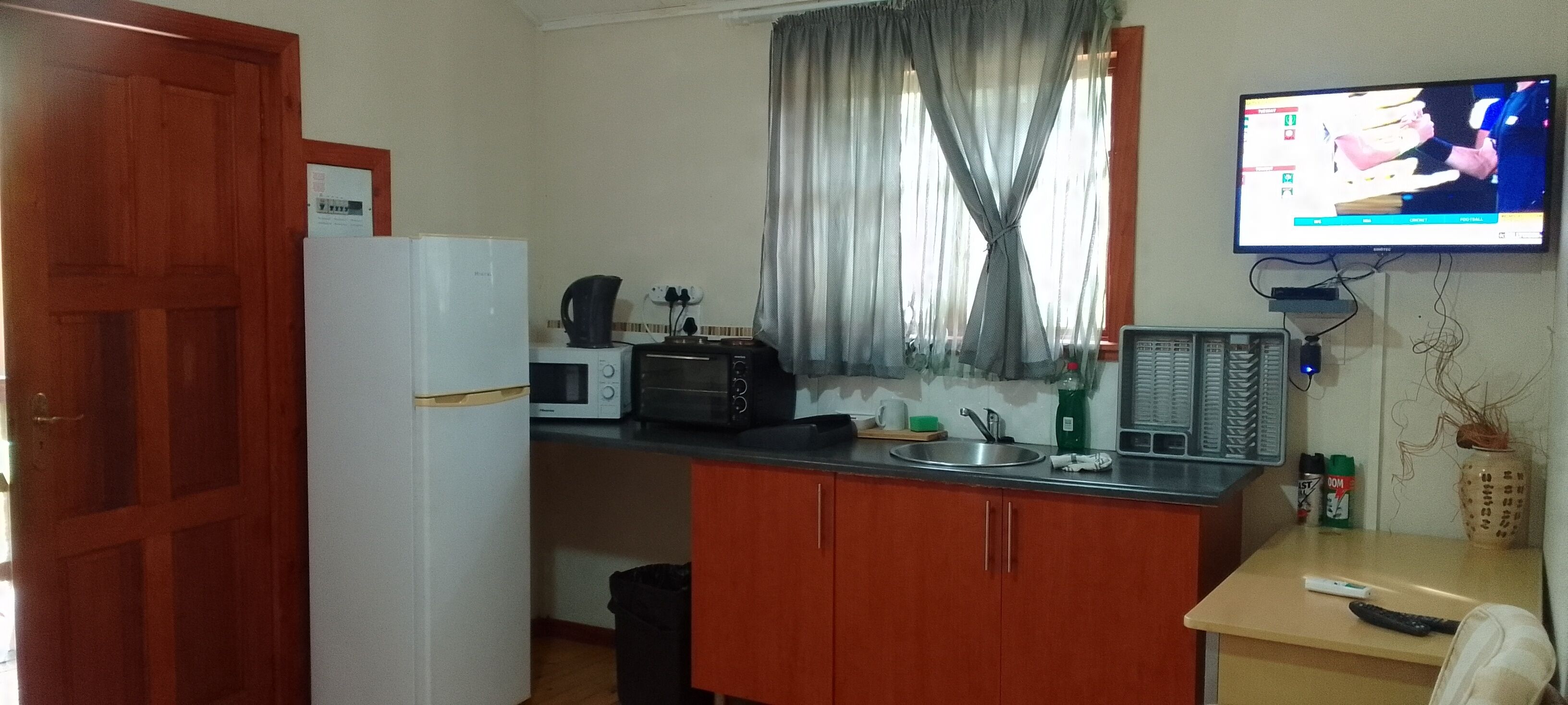 A compact kitchen with a refrigerator, microwave, toaster, and a sink. A television is mounted on the wall, and there is a small table with a remote control and a plant.