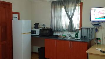 A compact kitchen with a refrigerator, microwave, toaster, and a sink. A television is mounted on the wall, and there is a small table with a remote control and a plant.