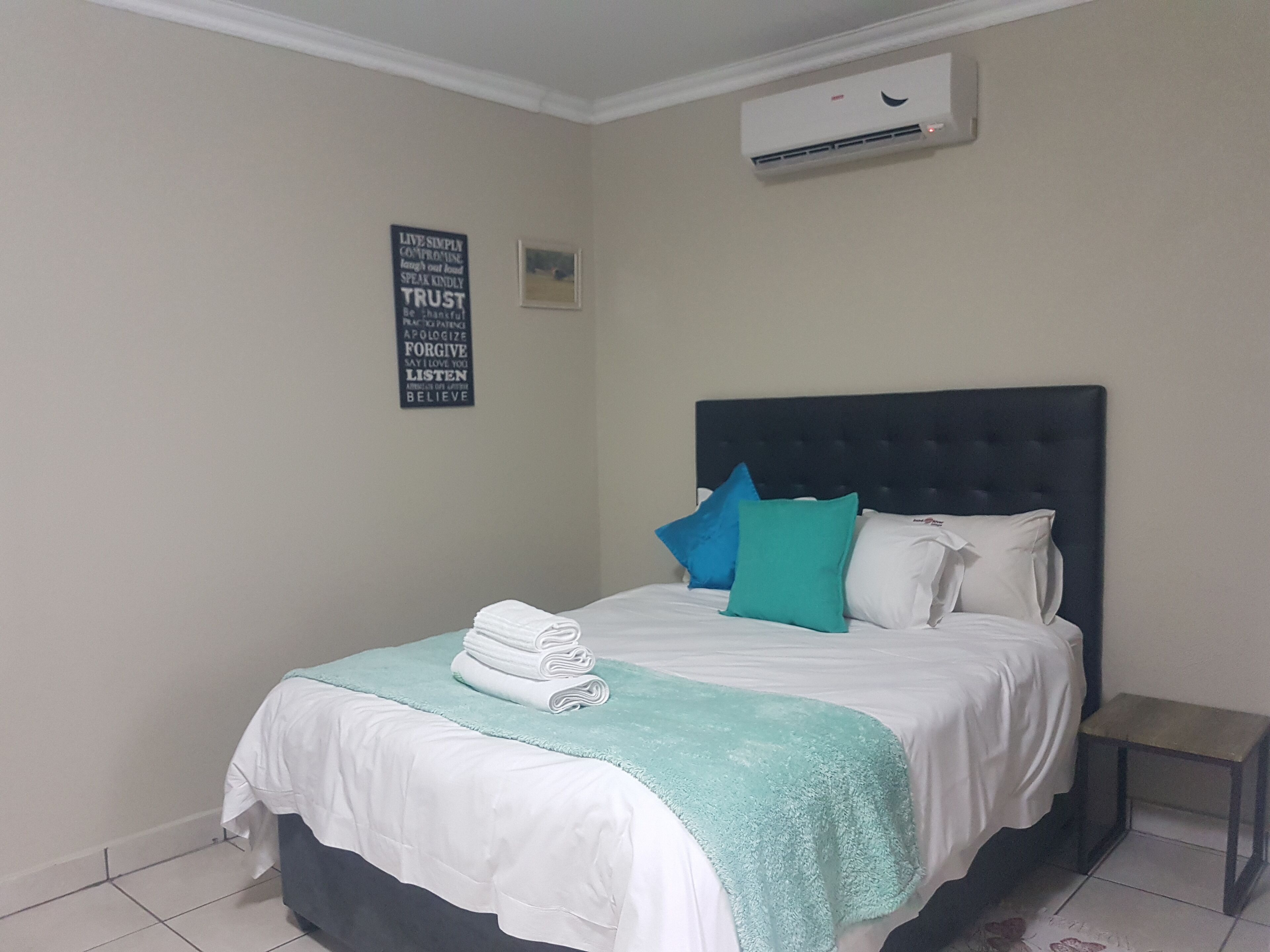 A neatly made bed with a teal throw blanket and pillows, a small bedside table, and a wall-mounted air conditioner.