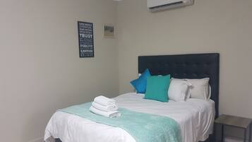 A neatly made bed with a teal throw blanket and pillows, a small bedside table, and a wall-mounted air conditioner.