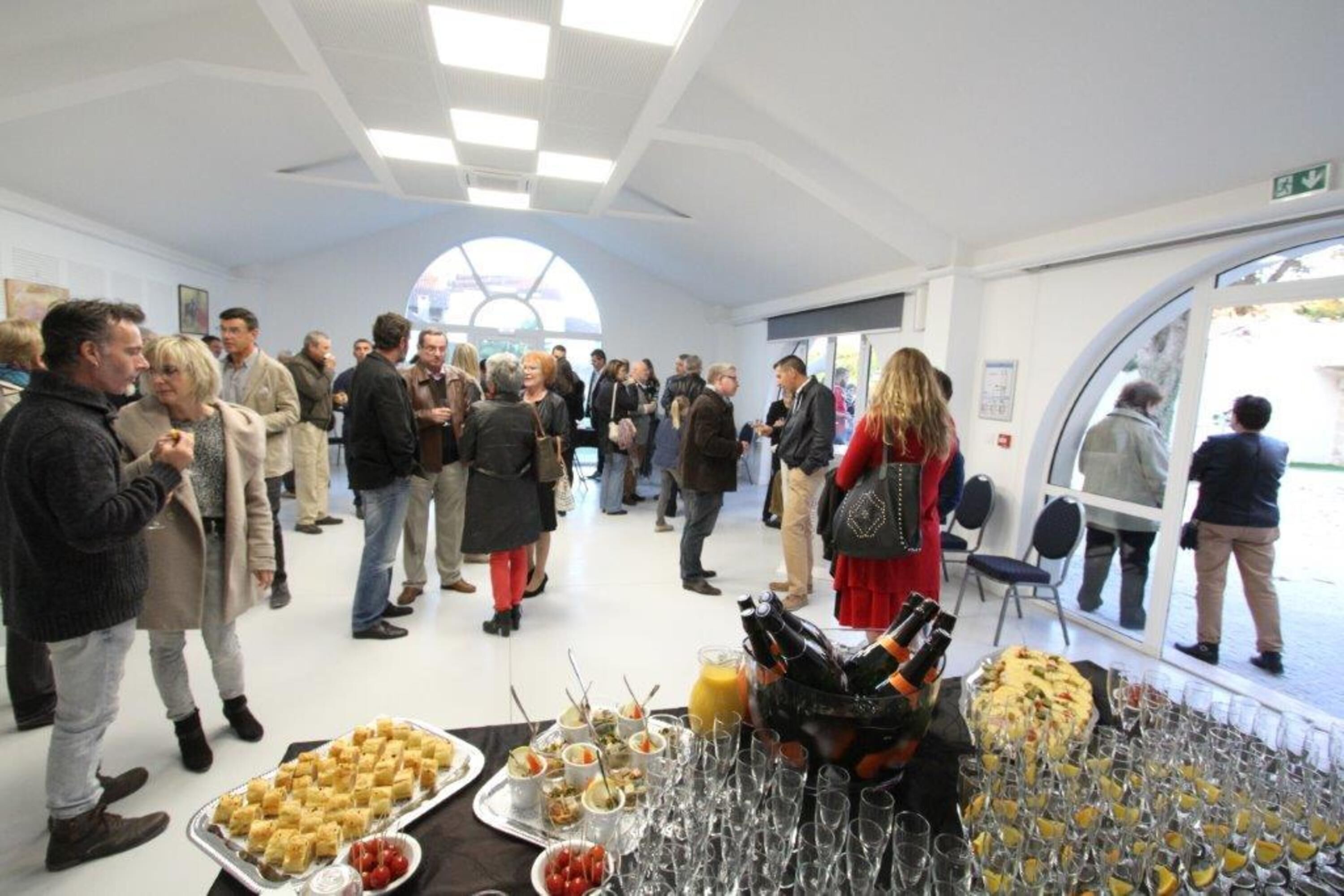 Un evento social con gente interactuando en un salón con techo alto y amplias ventanas.