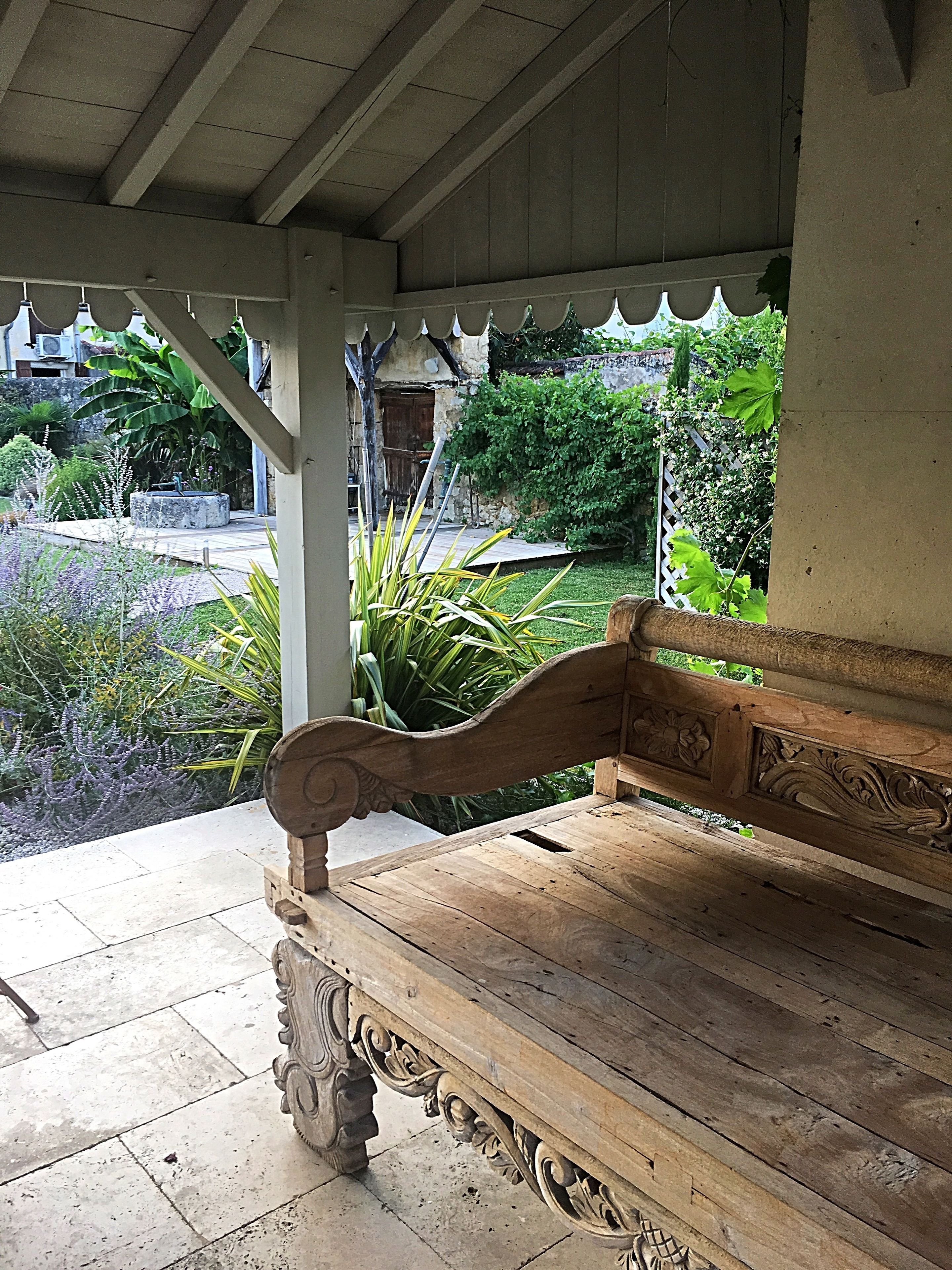 Un banc en bois orné de sculptures complexes, placé sous un porche abrité, d’où l’on peut admirer un jardin et, au loin, un bâtiment en pierre.