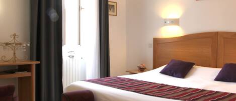 Una habitación de hotel con cama, escritorio, silla y ventana con cortinas.