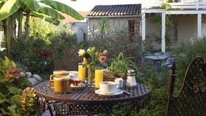 Une table ronde en métal, dressée pour le petit-déjeuner, entourée d’une végétation luxuriante et avec une maison en arrière-plan.