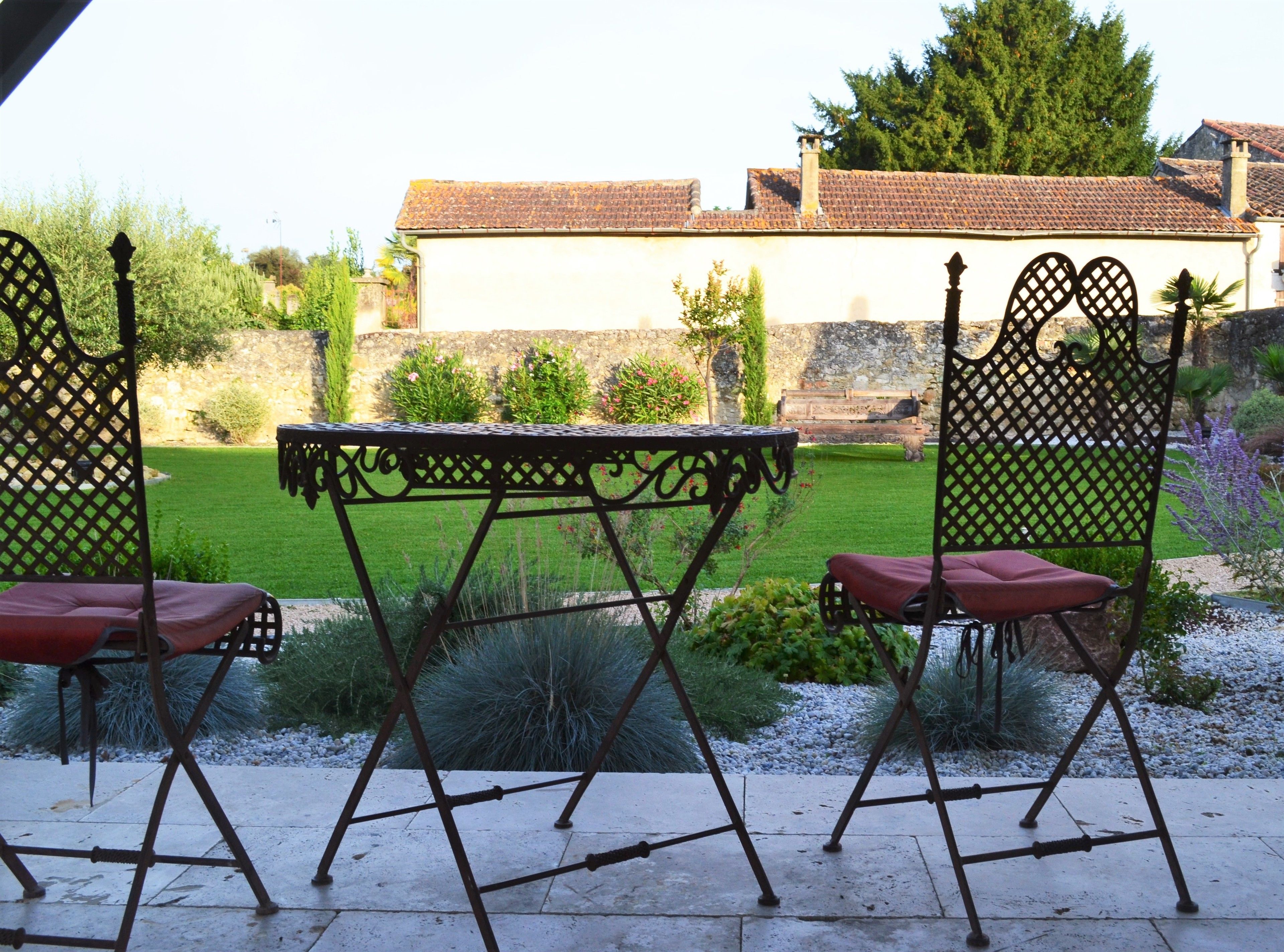 A patio with a metal table and chairs, surrounded by a garden with various plants and a stone wall in the background.