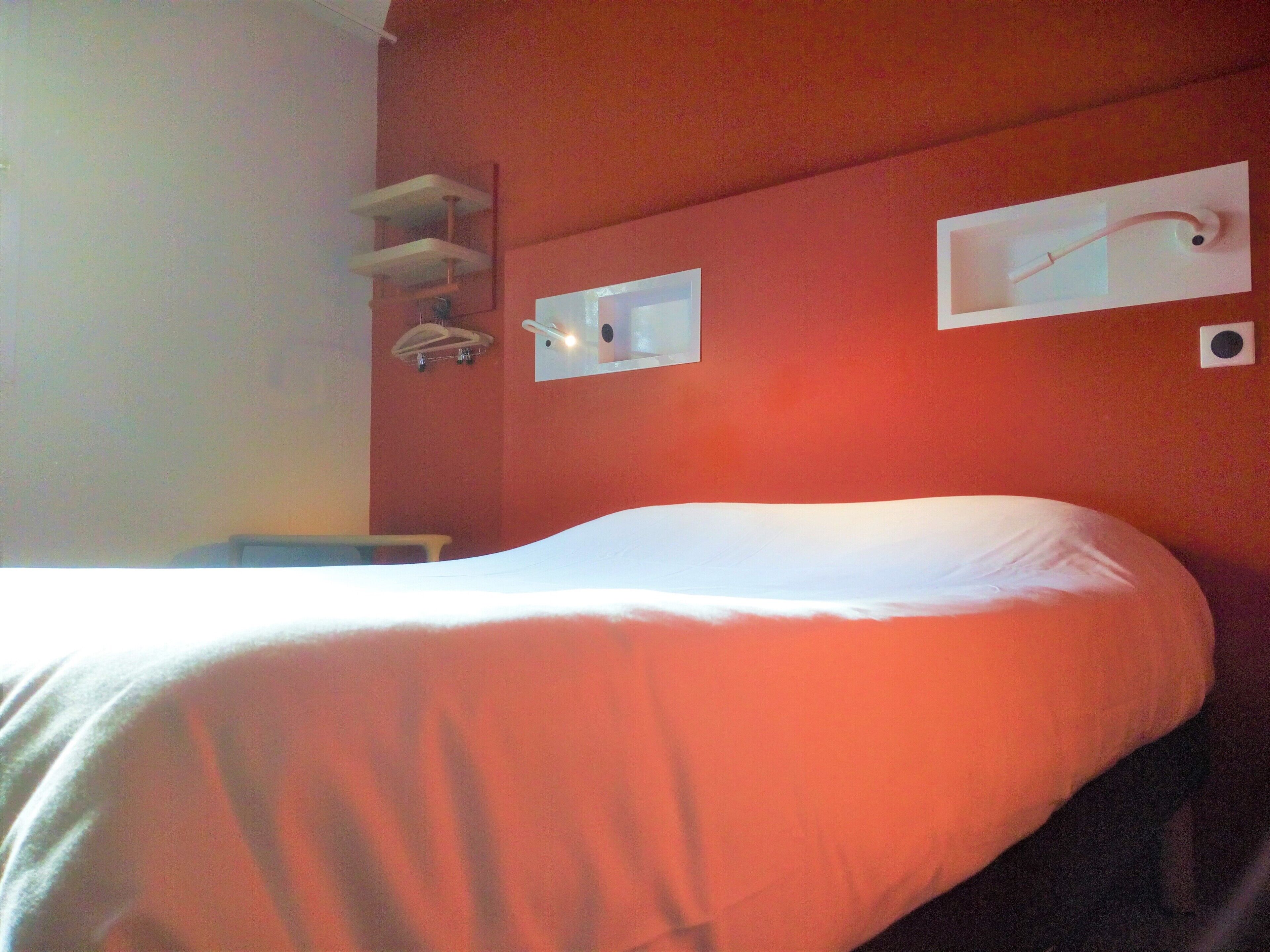Una camera d'albergo con un letto, una parete bianca e uno scaffale con un asciugamano.
