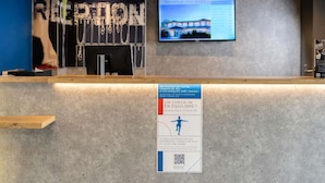 A reception desk with a monitor displaying a welcome message, a sign with a QR code, and a blue wall with a partial view of a mannequin. - Ibis Budget Perigueux Boulazac (Boulazac-Isle-Manoire)
