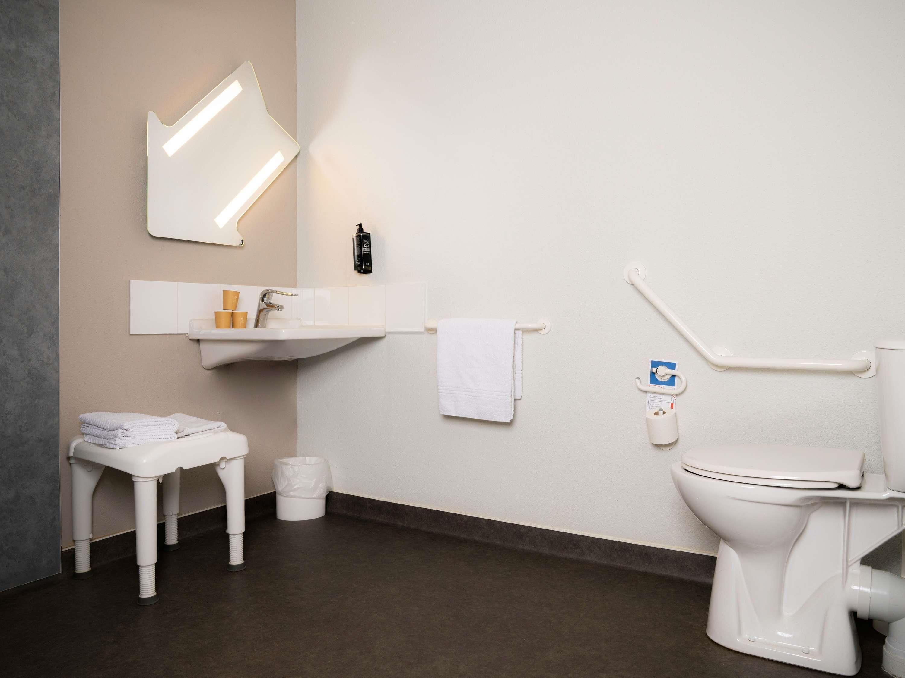 Un baño moderno con un lavamanos, espejo y retrete blancos. Hay un taburete pequeño y un cesto de basura.