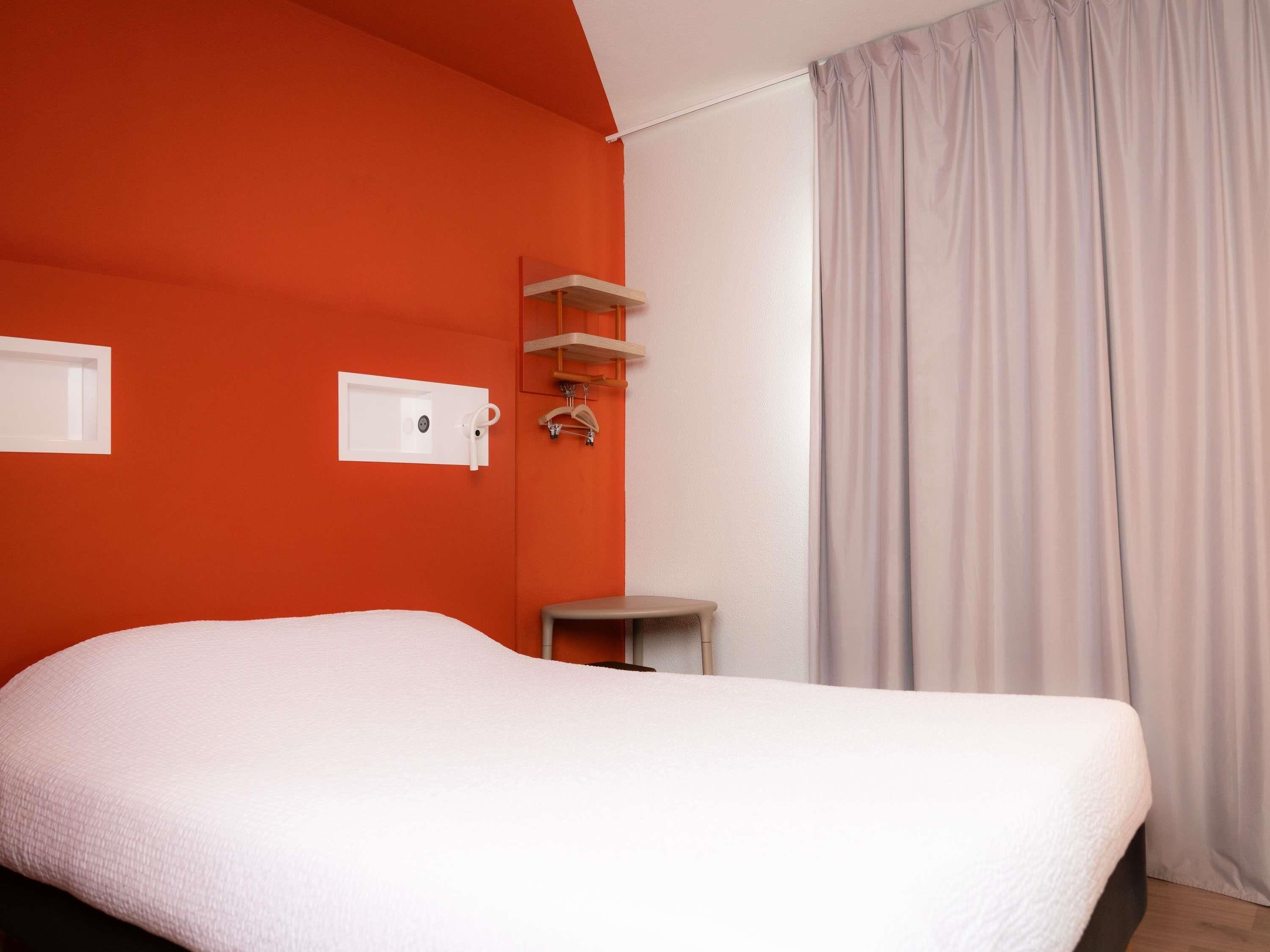 A hotel room with a bed, a small table, a shelf with hangers, and a window with curtains.