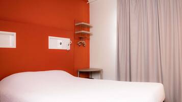 A hotel room with a bed, a small table, a shelf with hangers, and a window with curtains.