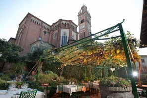 An outdoor dining area with tables and chairs, a pergola with climbing plants, and a historic building with a clock tower in the background.