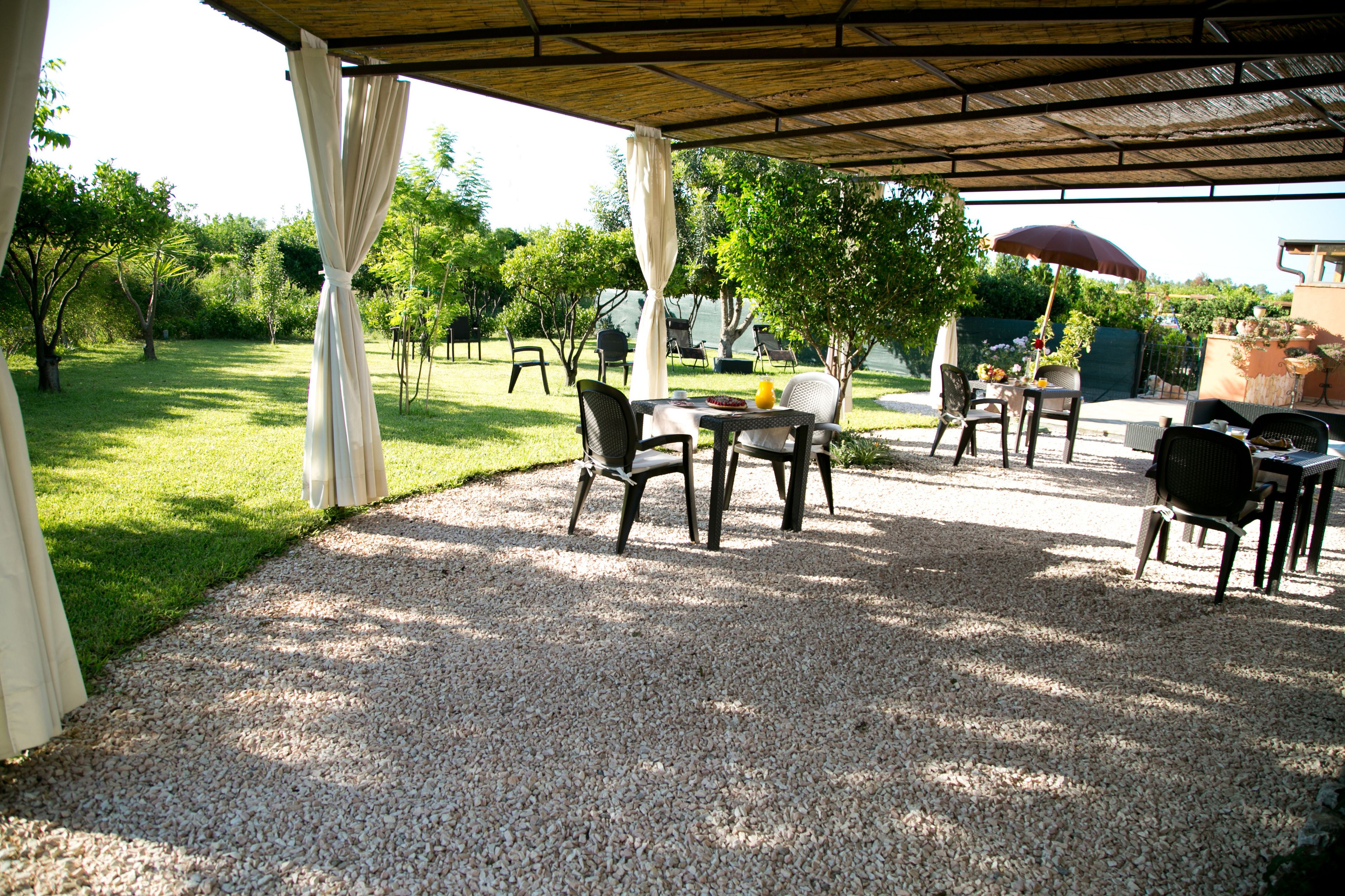 Eine Terrassengaststätte mit Tischen und Stühlen, ein überdachter Bereich mit Dach und Blick auf einen Garten mit Bäumen und einem Gebäude im Hintergrund.