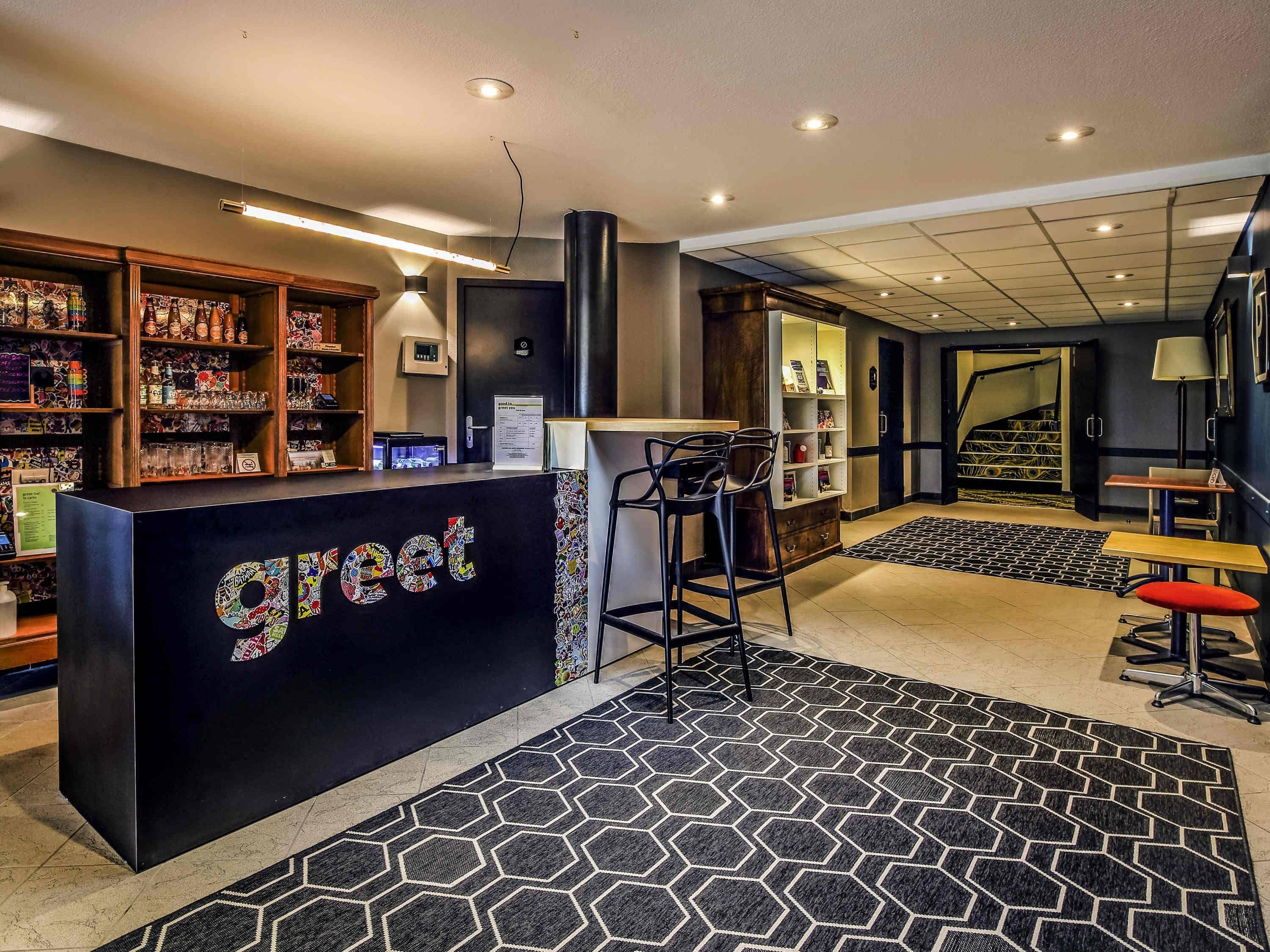 Un espace d’accueil d’hôtel avec un comptoir noir, des étagères en bois où sont exposés divers objets, un escalier et un tapis à motifs.