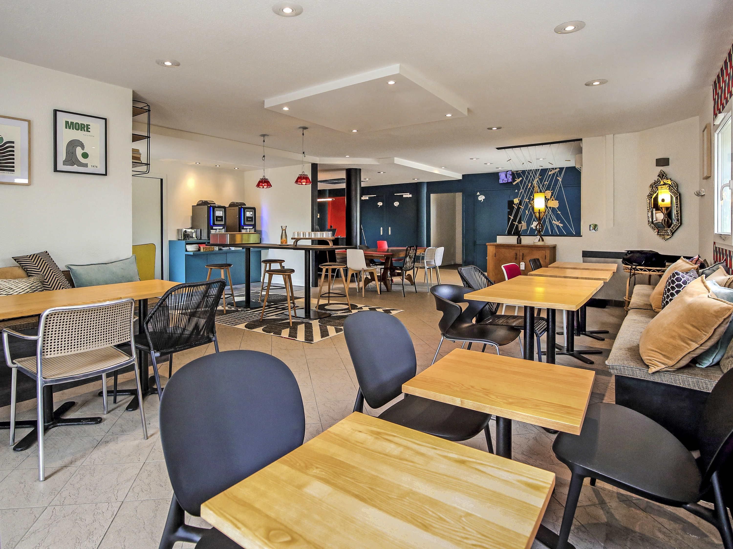 A modern cafe with wooden tables and black chairs, a bar area, and a seating area with sofas and cushions.