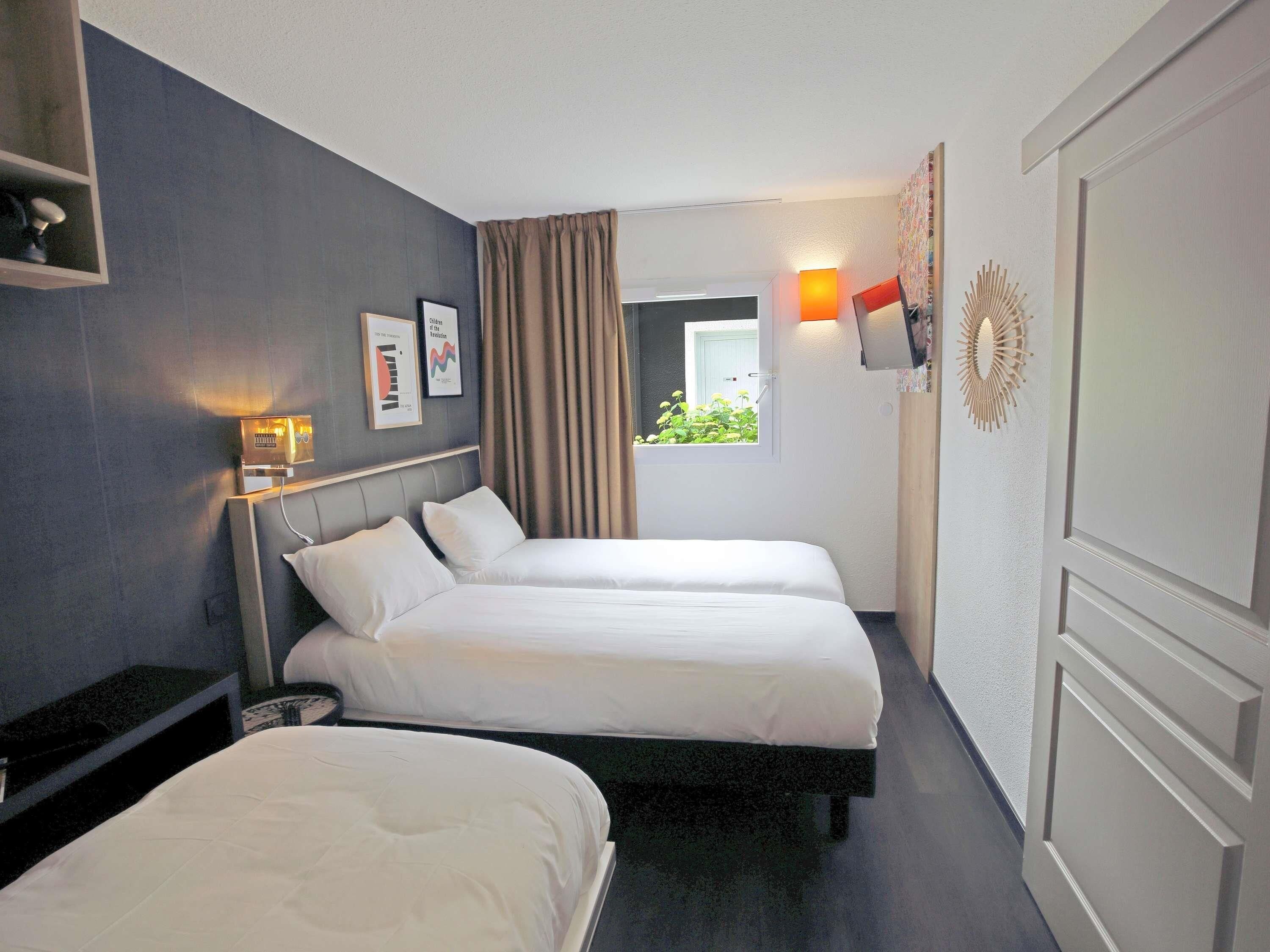 Une chambre d’hôtel avec deux lits, une fenêtre avec des rideaux, une lampe fixée au mur et une petite étagère avec quelques objets.