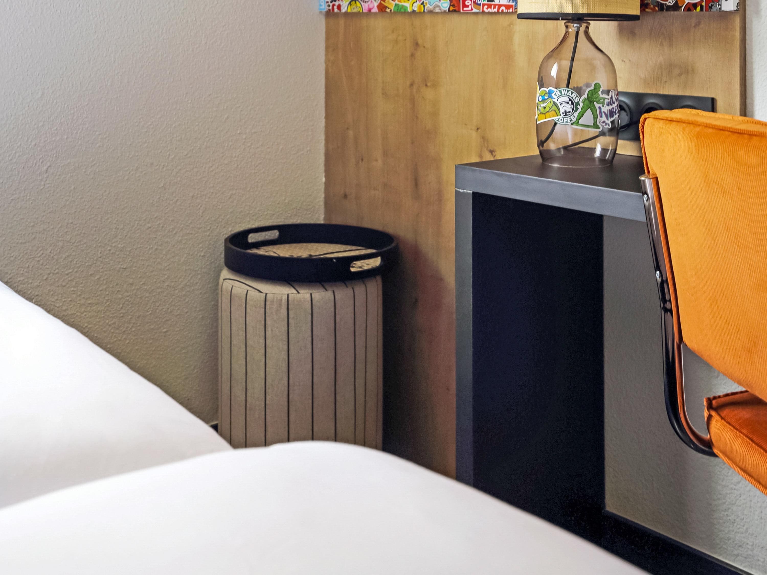 Una habitación de hotel con una cama, un pequeño bote de basura, un escritorio con una lámpara y una silla.