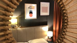 A circular mirror with a bamboo frame reflecting a bed, a wall with framed artwork, and a bedside lamp.