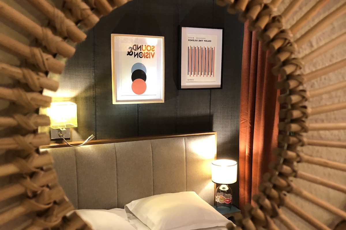 A circular mirror with a bamboo frame reflecting a bed, a wall with framed artwork, and a bedside lamp.