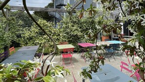 An outdoor seating area with tables and chairs, surrounded by greenery and a building with a 'greet' sign.