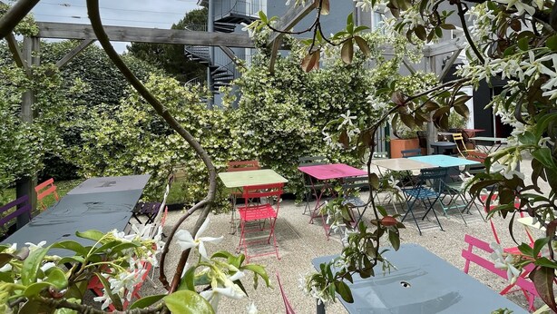 Een terras met tafels en stoelen, omringd door groen en een gebouw met een 'greet'-bord.