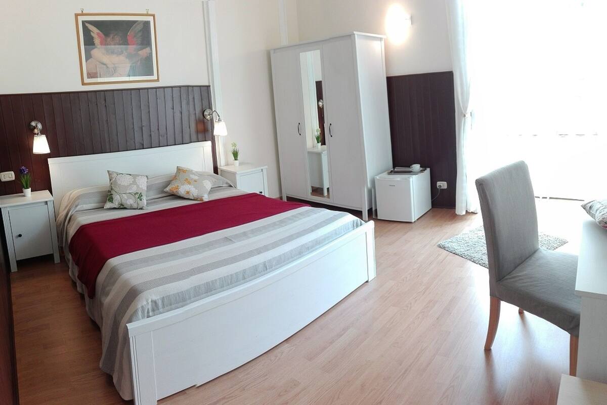 A modern hotel room with a large bed, a wardrobe, a desk, and a chair.