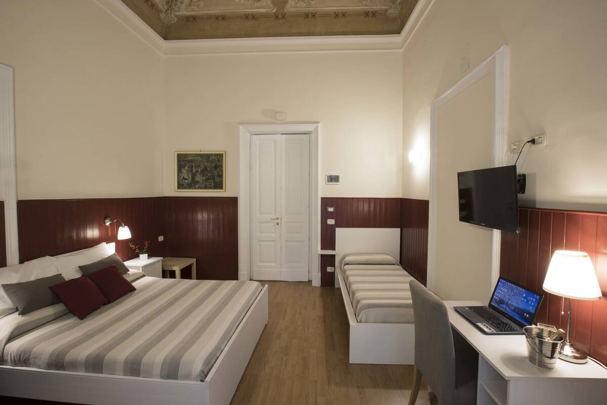 A hotel room with a bed, a desk with a laptop, a TV, and a bedside table with a lamp.