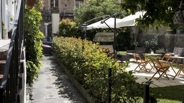 A narrow pathway flanked by greenery, leading to an outdoor seating area with tables and chairs under a white umbrella. - Dimora Novecento (Catania)