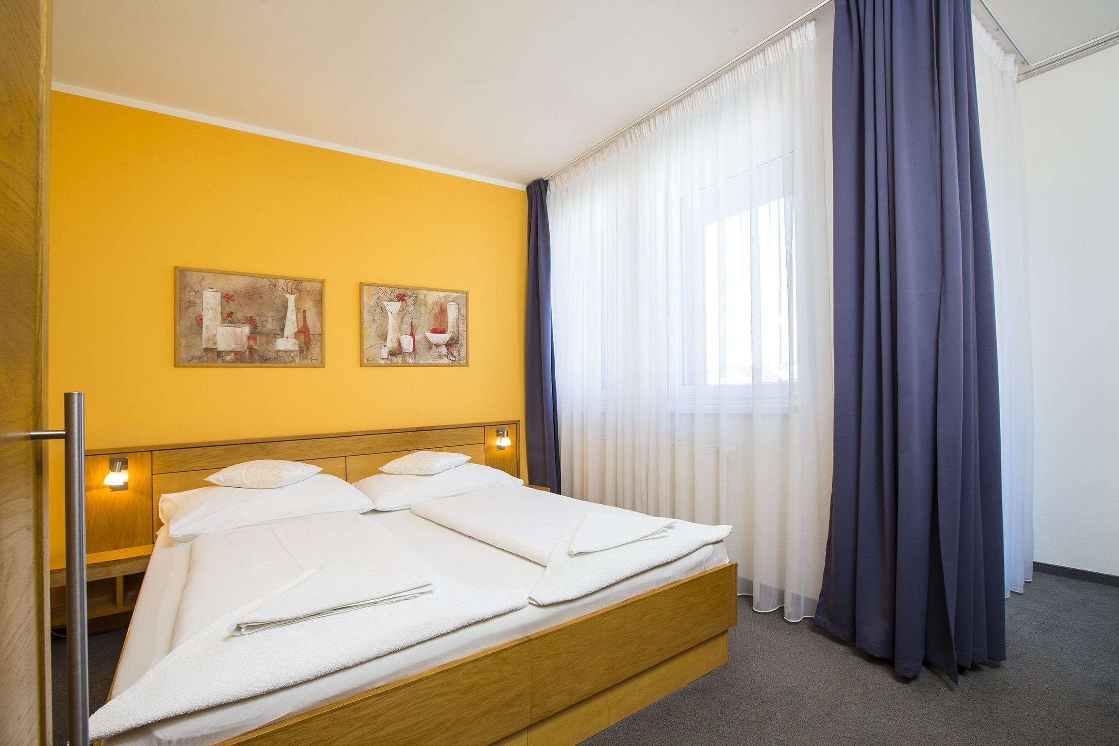 A hotel room with a bed, two paintings on the wall, and a window with curtains.