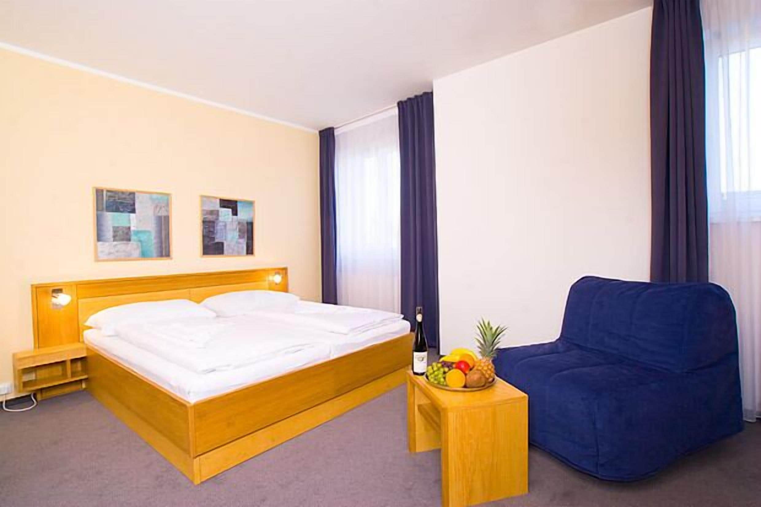 A hotel room with a wooden bed, a blue sofa, a small wooden side table with fruit, and two framed pictures on the wall.