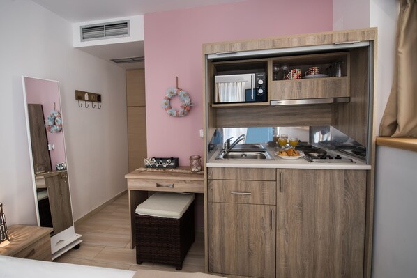 Cozinha compacta com micro-ondas, pia e bancada pequena. Há um banquinho de vime e um porta-roupas fixado na parede.