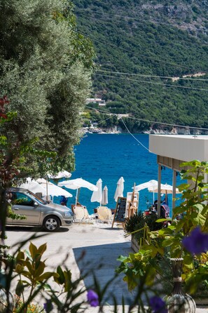 A coastal view with a car parked near a building with outdoor seating and umbrellas, overlooking the sea. - Ilianthos Apartments & Studios (Lefkada)